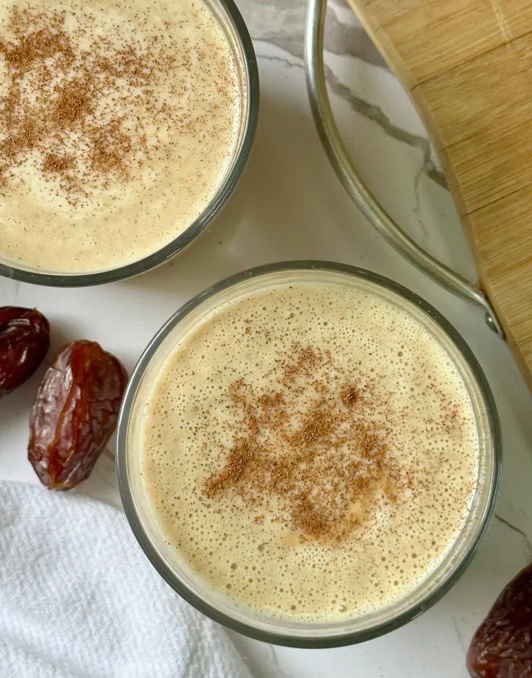 2 glasses of yellow banana date smoothie with cinnamon sprinkled on top and dates and a cutting board in the background