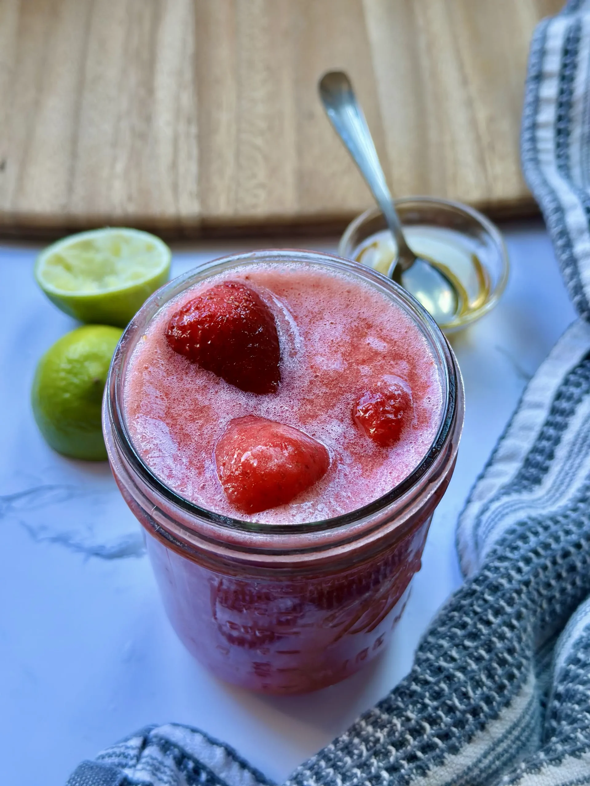 strawberry smoothie sitting on a white counter with squeezed lime and a small bowl of honey in the background