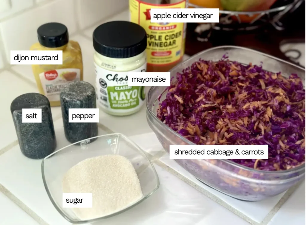 purple cabbage coleslaw ingredients sitting on a counter including a big tupperware of shredded cabbage and carrots, a small bowl of sugar, salt and pepper shakers, and a jar of mayonnaise, a jar of apple cider vinegar and a jar of mustard