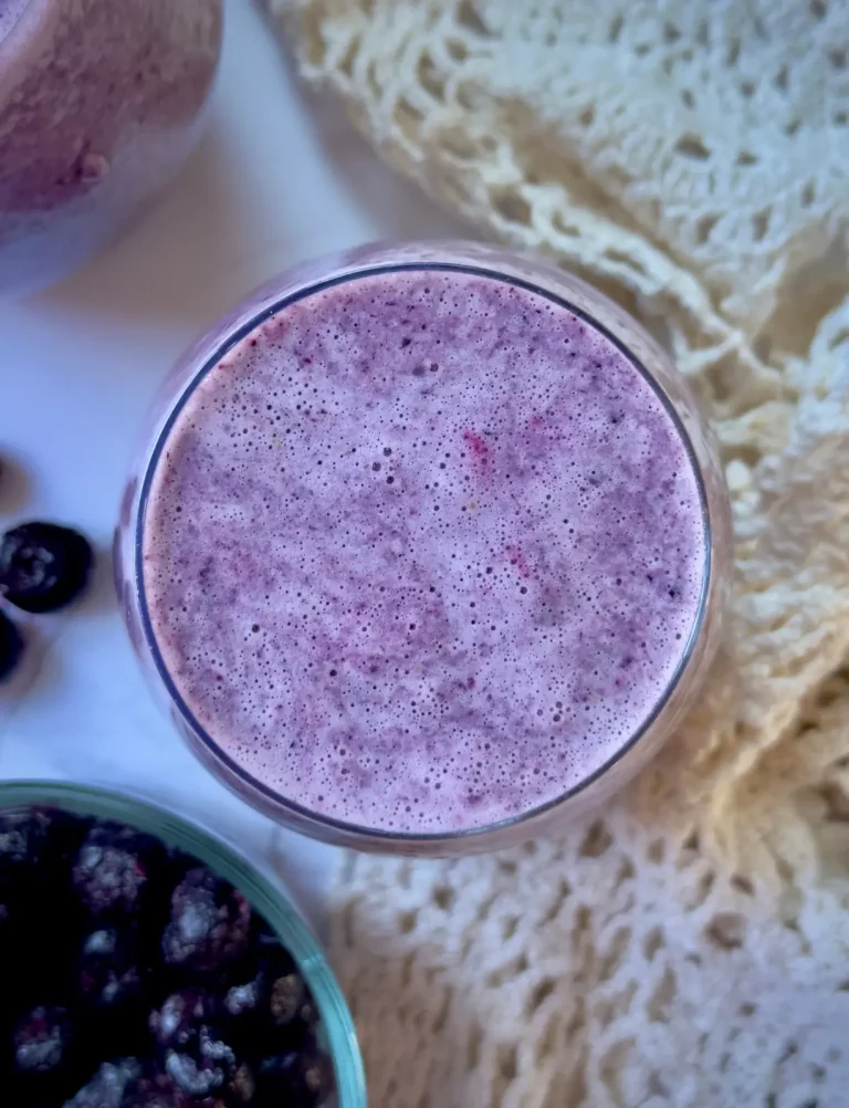 3 ingredient blueberry smoothie feature image of a purple blueberry smoothie with a doily and a glass bowl of blueberries in the background