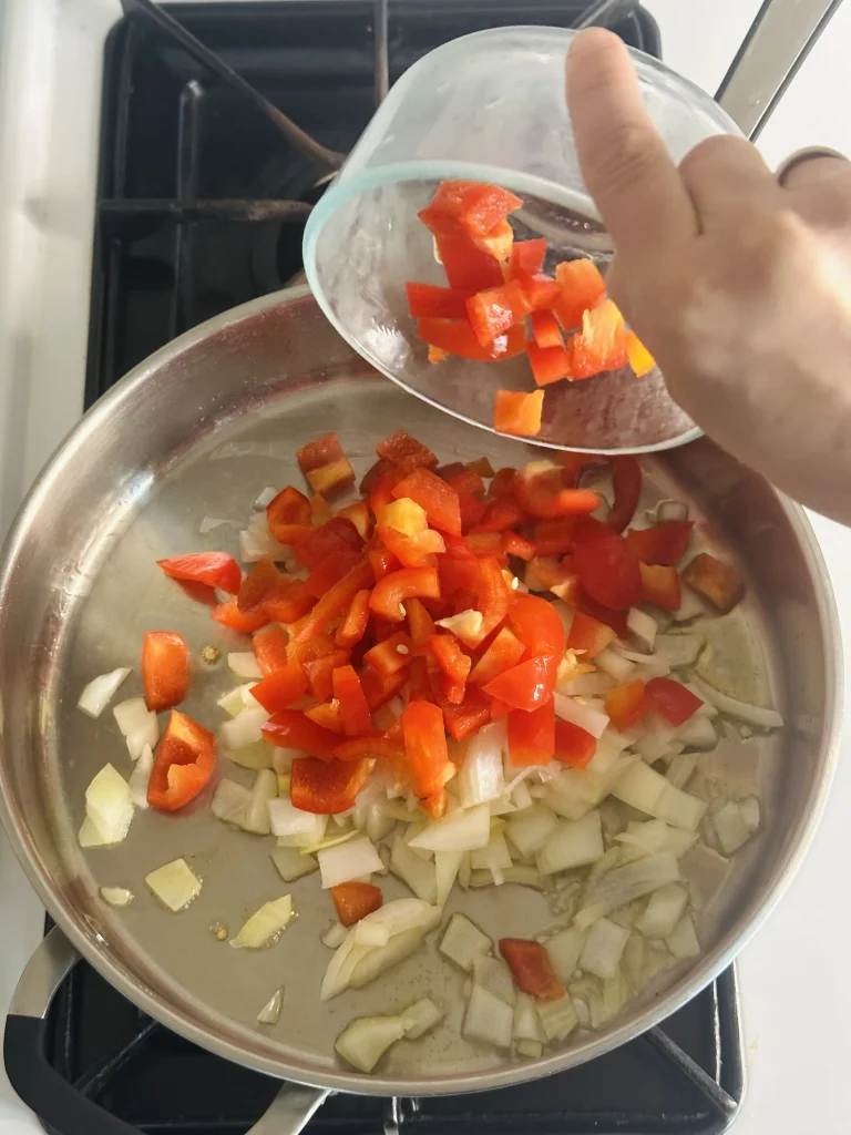 pouring red bell peppers from a glass bowl into a pan of onions