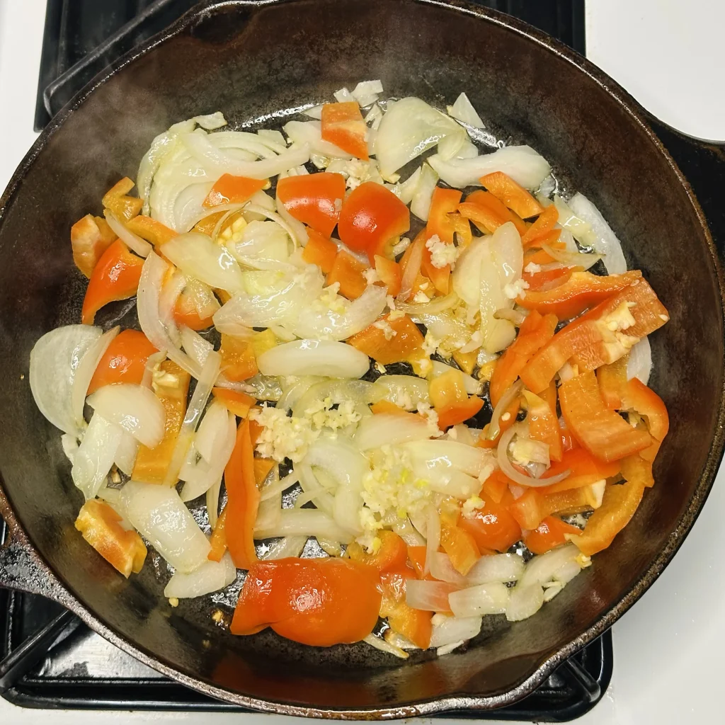 adding minced garlic to onion and red bell pepper cooking in a cast iron pot for tofu and broccoli stir fry