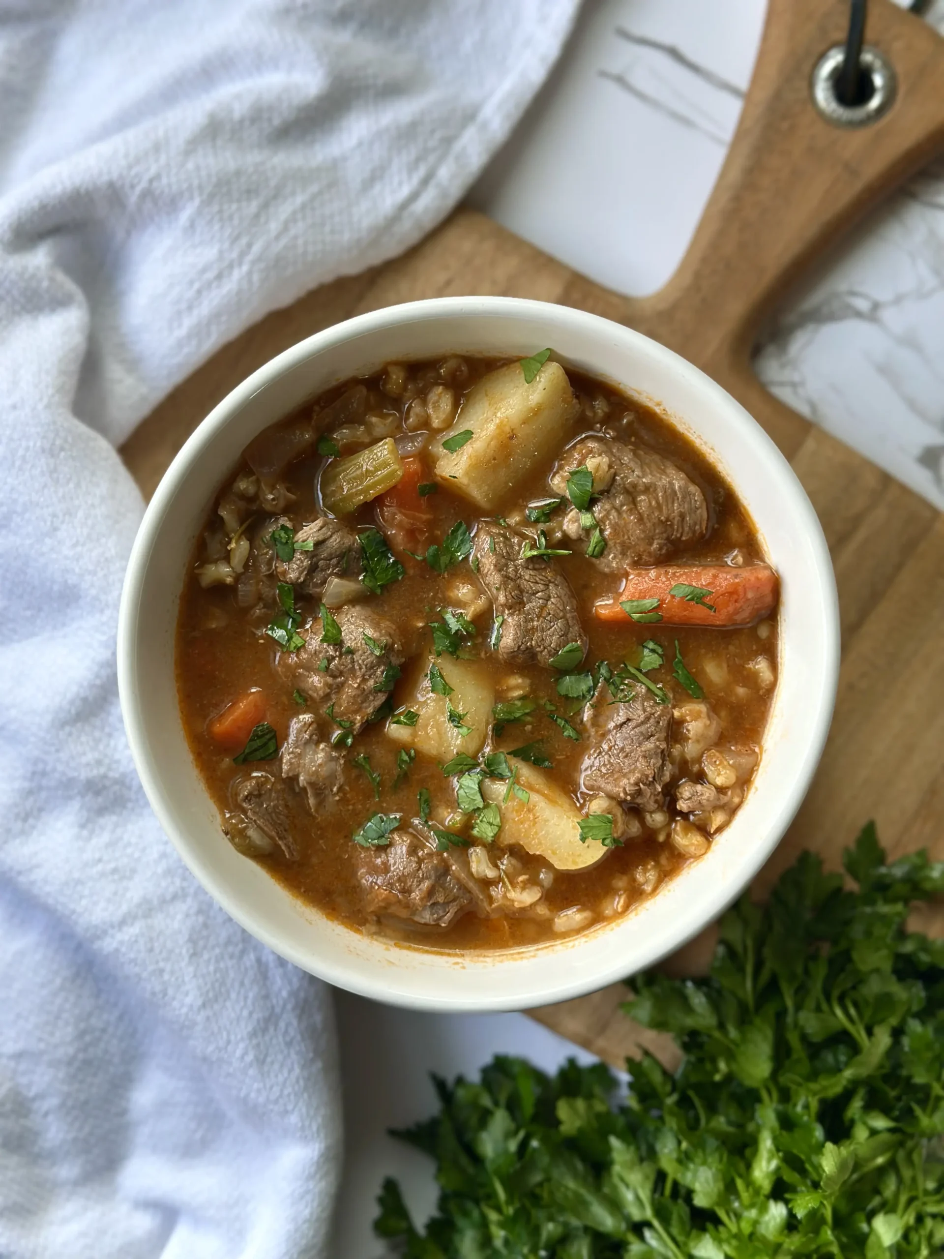 beef and barley vegetable soup in a white bowl sprinkled with parsley, sitting on a wooden cutting board with a white napkin and a bunch of fresh parsley in the background