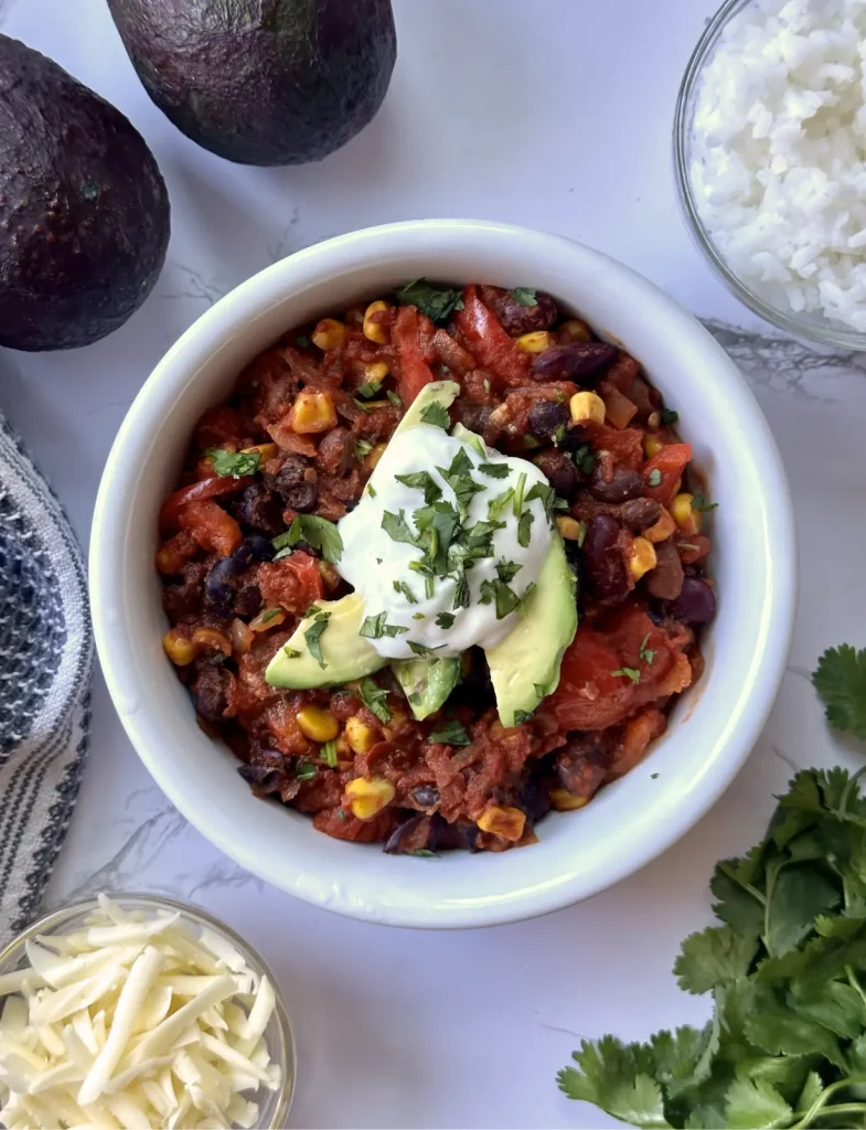 black bean chili with corn feature image with a white bowl of red chili with sour cream and cilantro and avocado on top on a white counter with cilantro, rice and cheese in the background