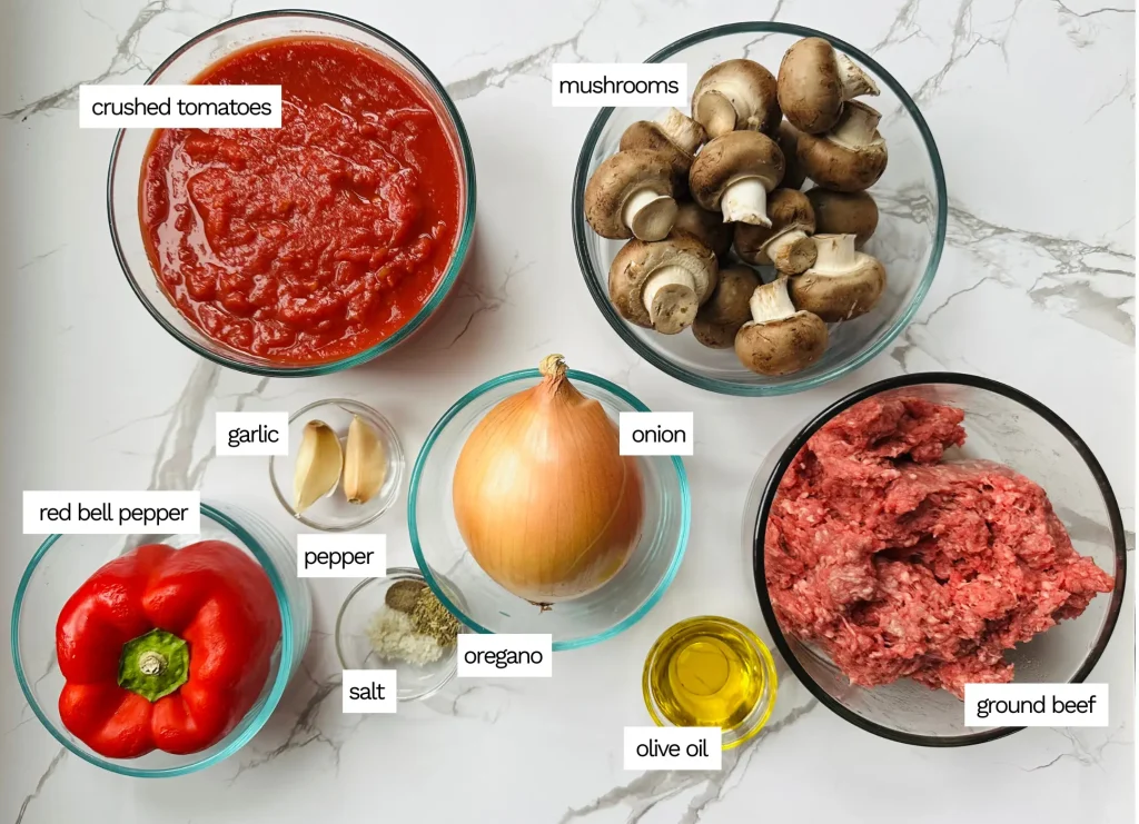 ingredients for meat sauce with mushrooms including mushrooms, crushed tomatoes, ground beef and seasonings on a white counter in glass bowls