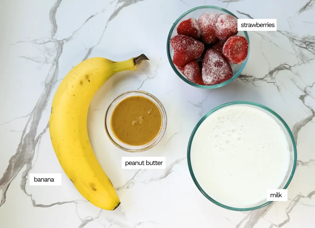 strawberry banana peanut butter smoothie ingredients including banana, strawberries, milk and peanut butter on a white marble counter