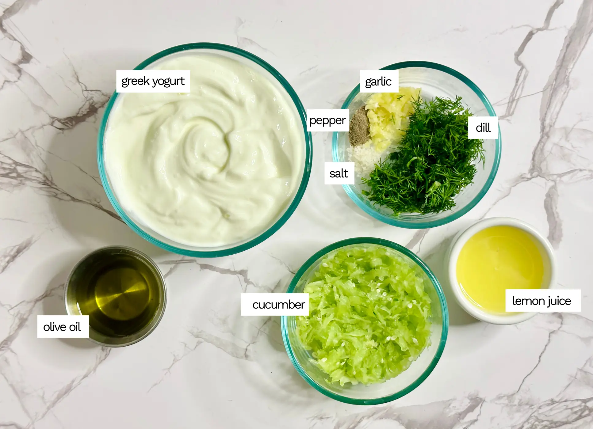 tzatziki ingredients including yogurt, dill, lemon juice and olive oil in small glass bowls on a white counter