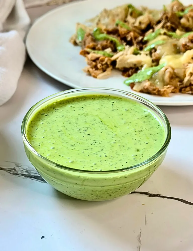 glass bowl of green cilantro yogurt sauce with a plate of nachos in the background