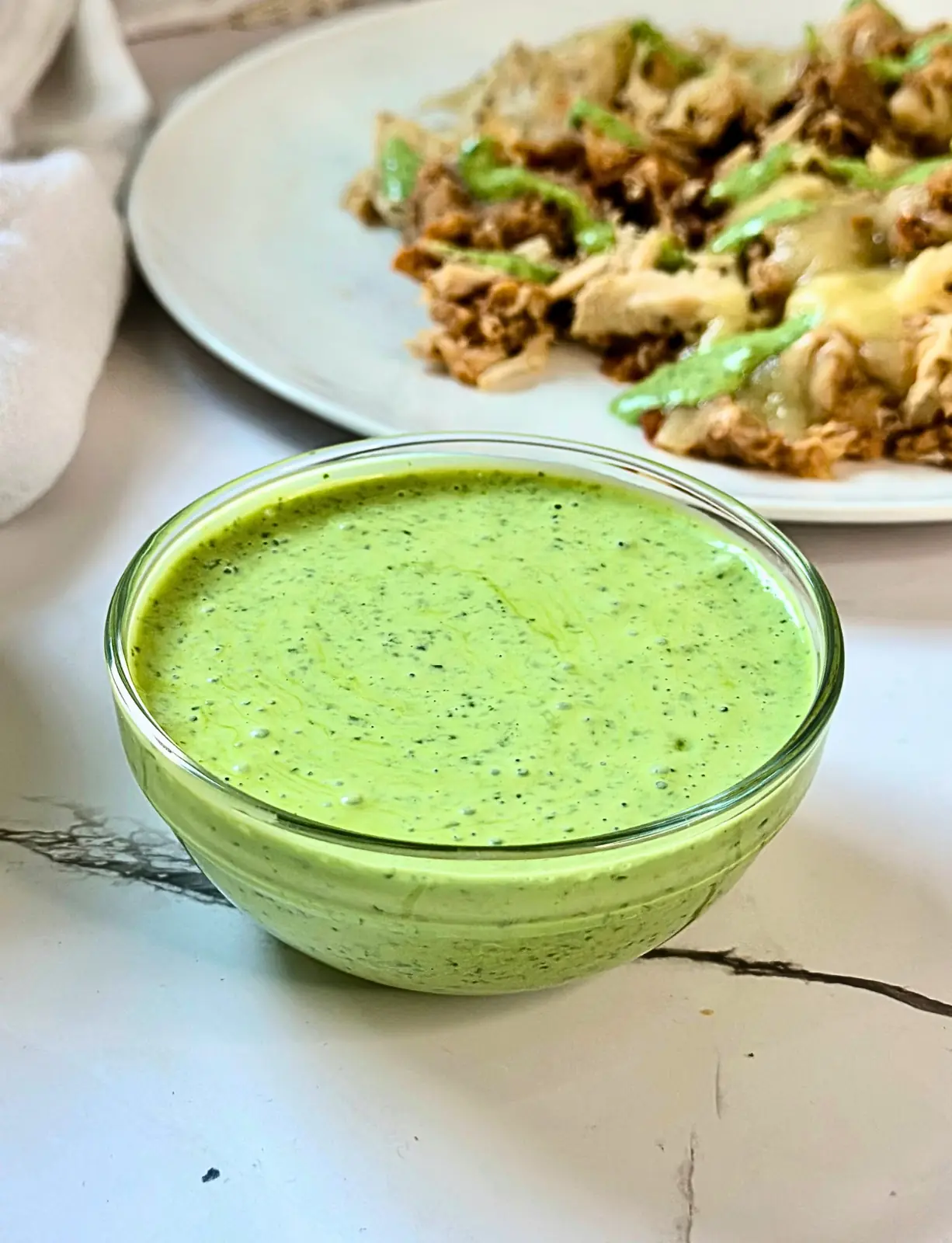 glass bowl of green cilantro yogurt sauce with a plate of nachos in the background